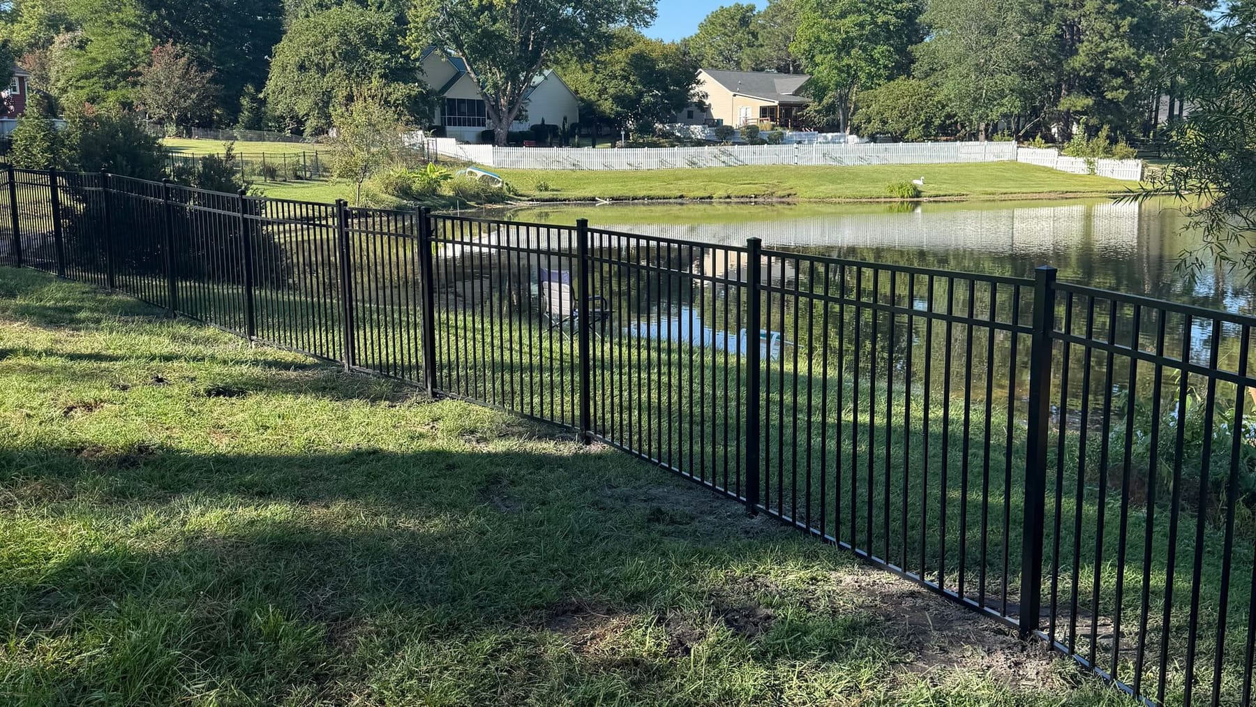 Black aluminum fence along a grassy pond bank, with homes across the water